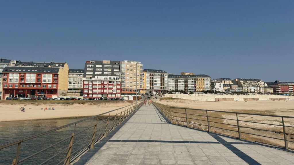 Pasarela de madera del paseo marítimo de A Rapadoira con playa a ambos lados y edificios de Foz al fondo