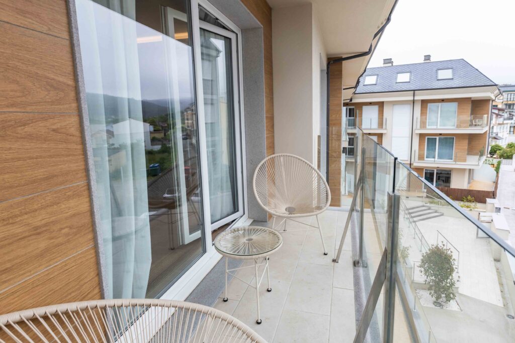 Terraza con sillas de mimbre blanco y barandilla de cristal en un apartamento Punta dos Prados de Apartagal